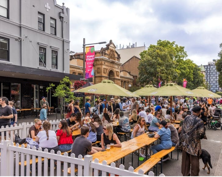 A wide shot of an outdoor street full of lots of people, with the main focus on the people siting at alfresco tables, chatting and drinking.