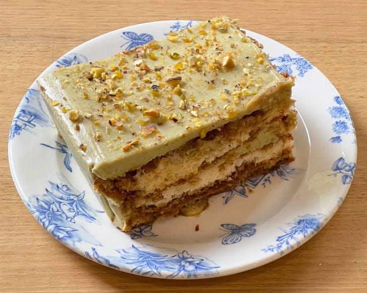 A close up of a slice of pistachio tiramisu presented on a decorative white plate. The pistachio tiramisu is a light green colour with a light sprinkle of crushed pistachio nuts on top.