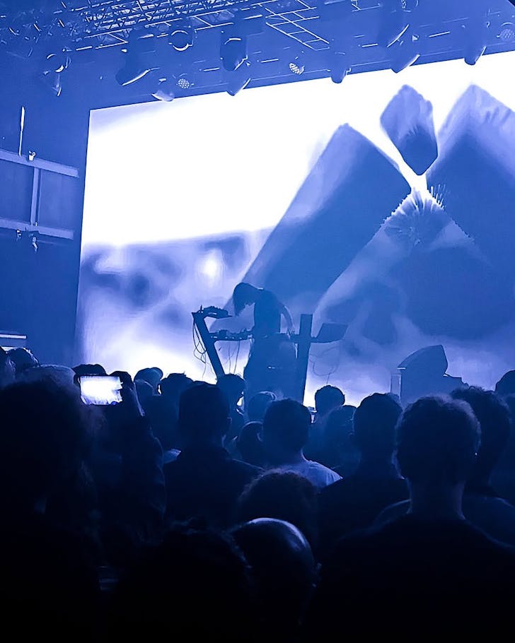 A moody, dark shot of Rival Consoles playing live in front of a purplely-blue A/V show.
