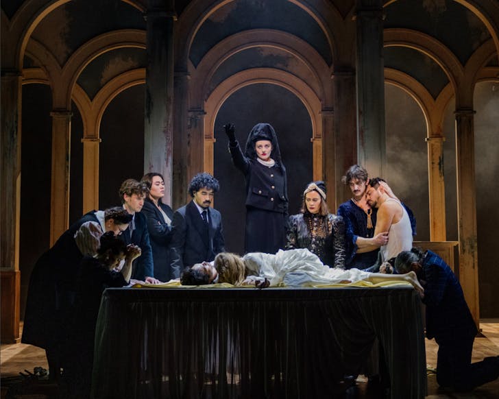 In a hall of arches, the cast of Romeo & Juliet crowd around the eponymous couple, who lie, motionless, on a table.