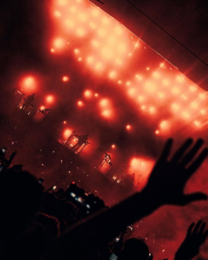 An angled shot of a silhouetted, orange-lighted crowd witnesses RÃFÃS DU SOL live onstage in Houston.