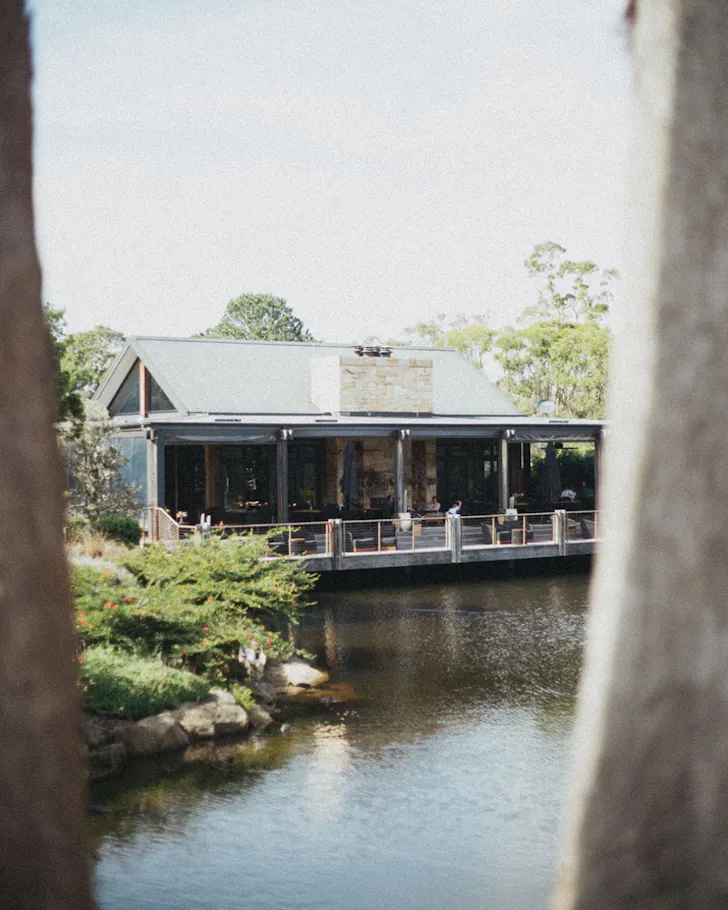 Waterside restaurant with a timber deck overlooking a tranquil lagoon, framed by trees and landscaped gardens on the Central Coast.