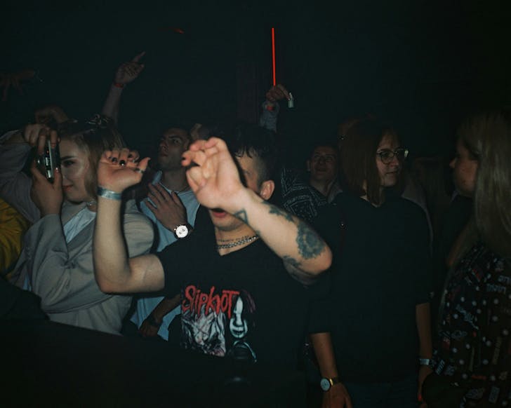 A vintage-90s-looking grainy photo of ravers in a dark venue, someone takes a photo with a point-and-shoot film camera.