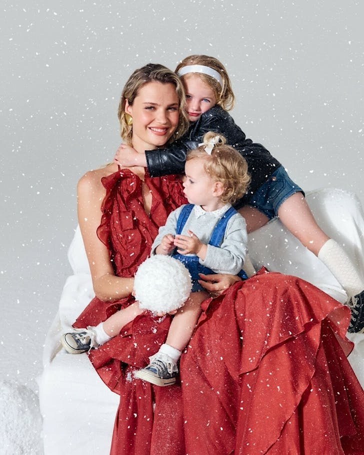 A woman in red and two children pose on a white chair with a fluffy snowball while it fake snows.
