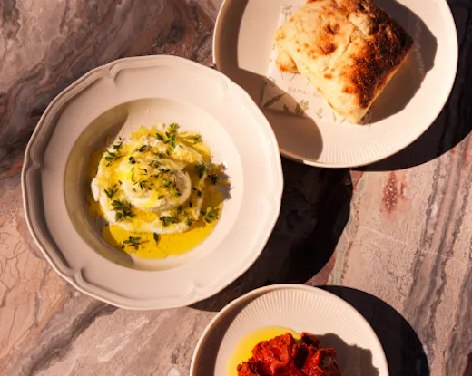Overhead view of three plated dishes on a pink marble table, including a creamy herb-topped dish, fresh bread and roasted red vegetables.