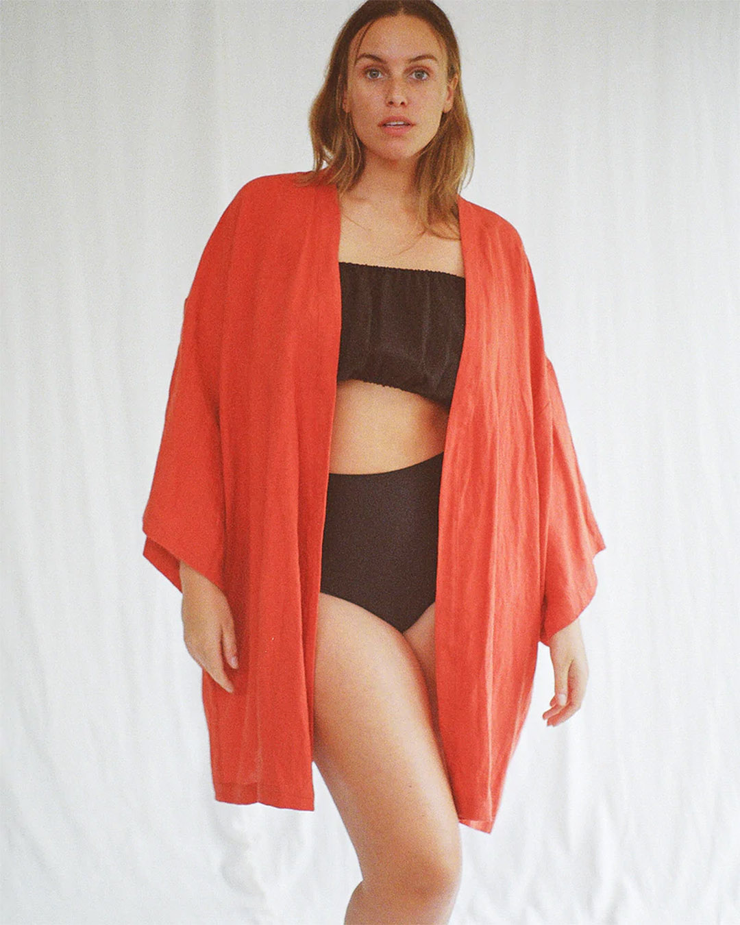 A woman wearing an orange kimono-style dress over brown high-waisted separates. 