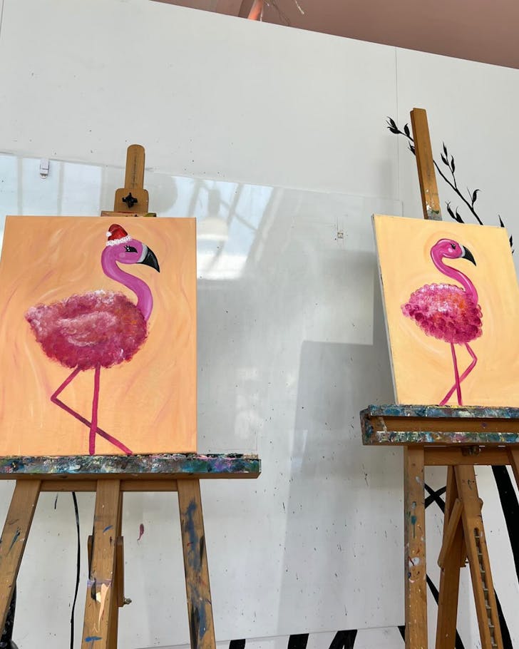 Two paintings of a falmingo on an orange background, both on easels, courtesy of a sip & paint session.