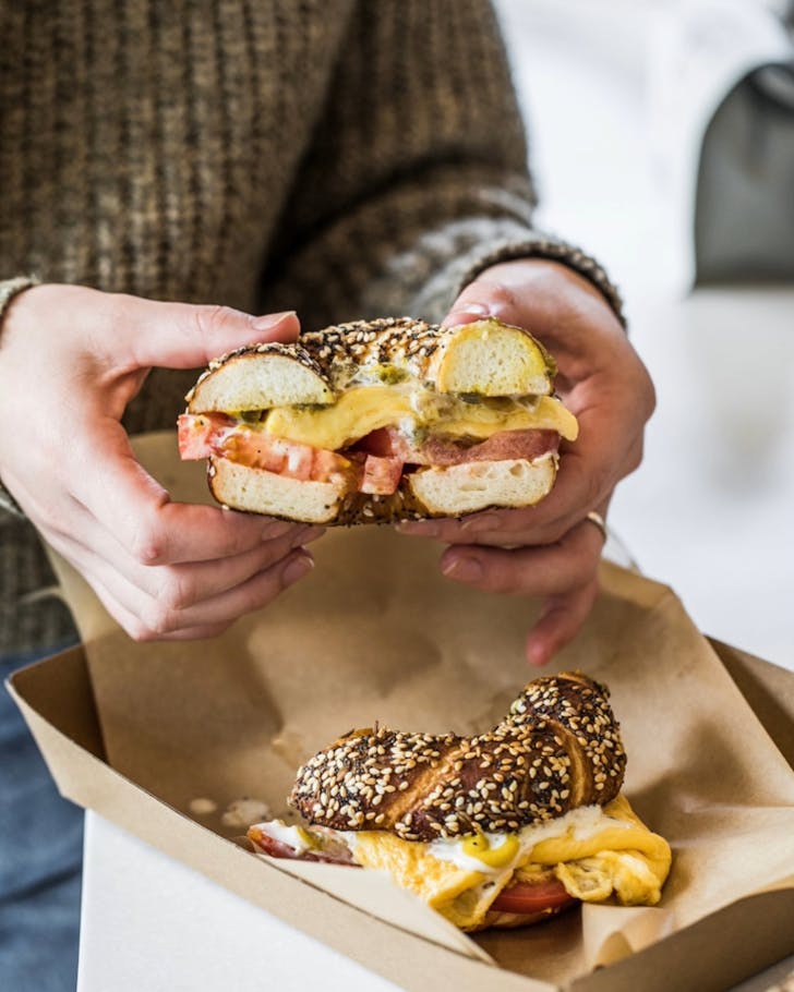 A close up image of two hands holding one half of a breakfast bagel, ready to eat, while the other half sits oozing out cheese on the table.