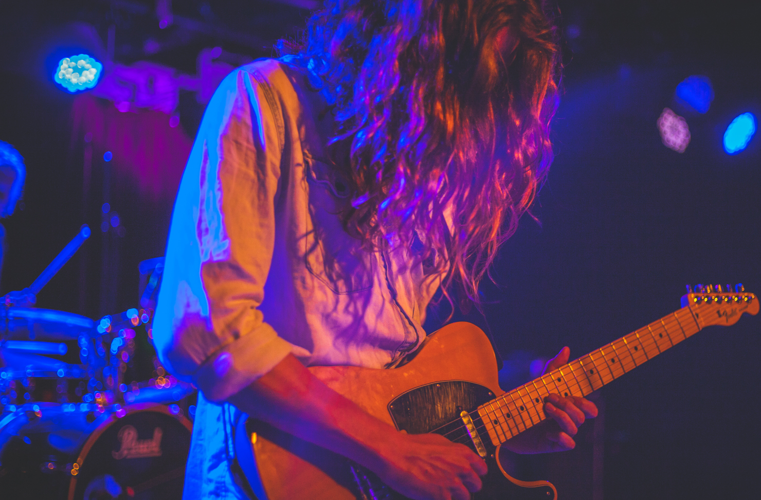 A musician on stage and playing guitar. Their face is covered by long, unruly hair. 