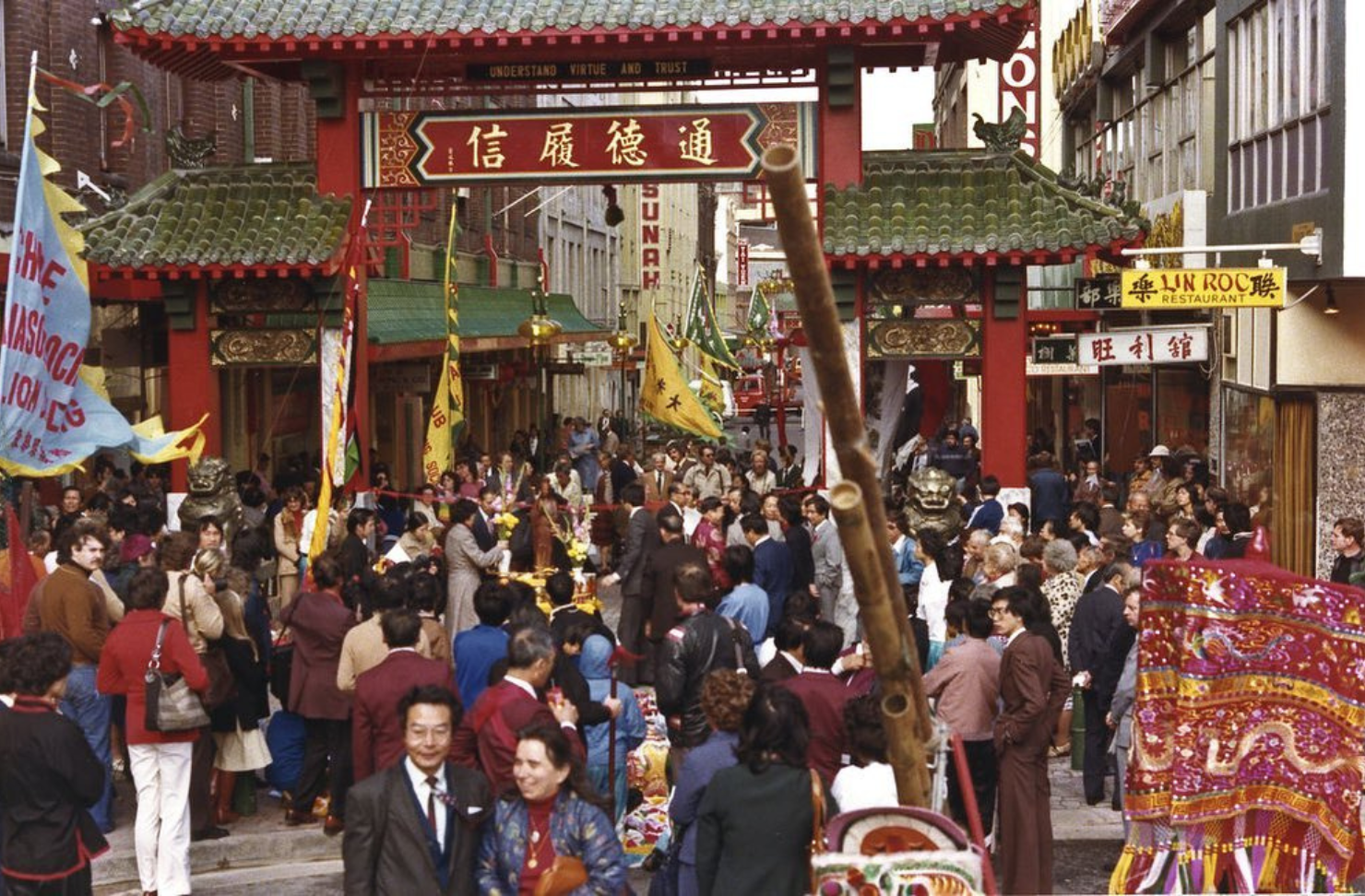 A bustling crowd gathers beneath Sydney Chinatown's ceremonial gate during a cultural festival, with lion dance performers, banners and spectators filling Dixon Street.