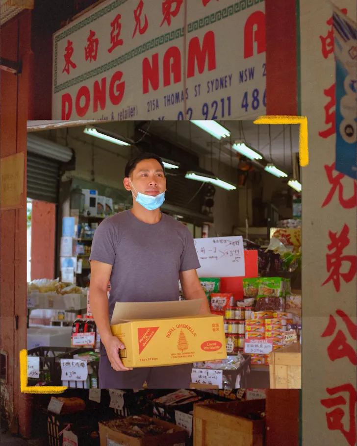 A shopkeeper stands at the entrance of Dong Nam grocery in Sydney's Chinatown holding a box of jasmine rice, surrounded by shelves of Asian pantry staples beneath the store's iconic red-and-white signage.