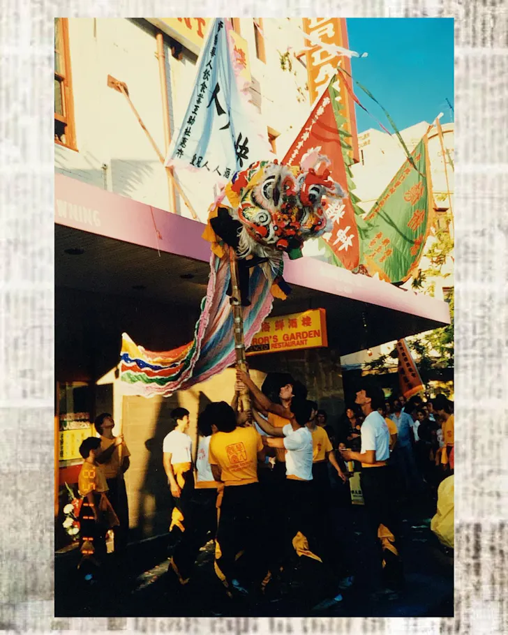 A traditional Chinese lion dance winds through Sydney's Chinatown during a street celebration, with performers carrying colourful flags as crowds gather outside local shopfronts.