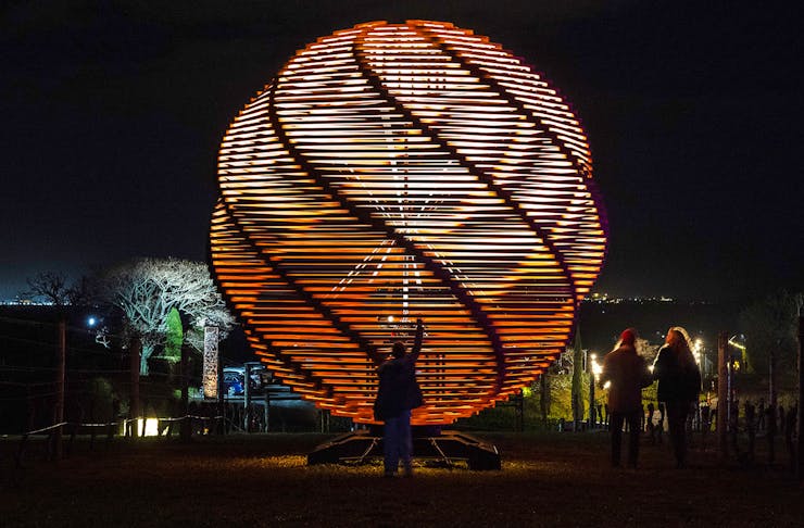 A silhouetted person stands in front of a giant wooden orb sculpture lit from the inside. 