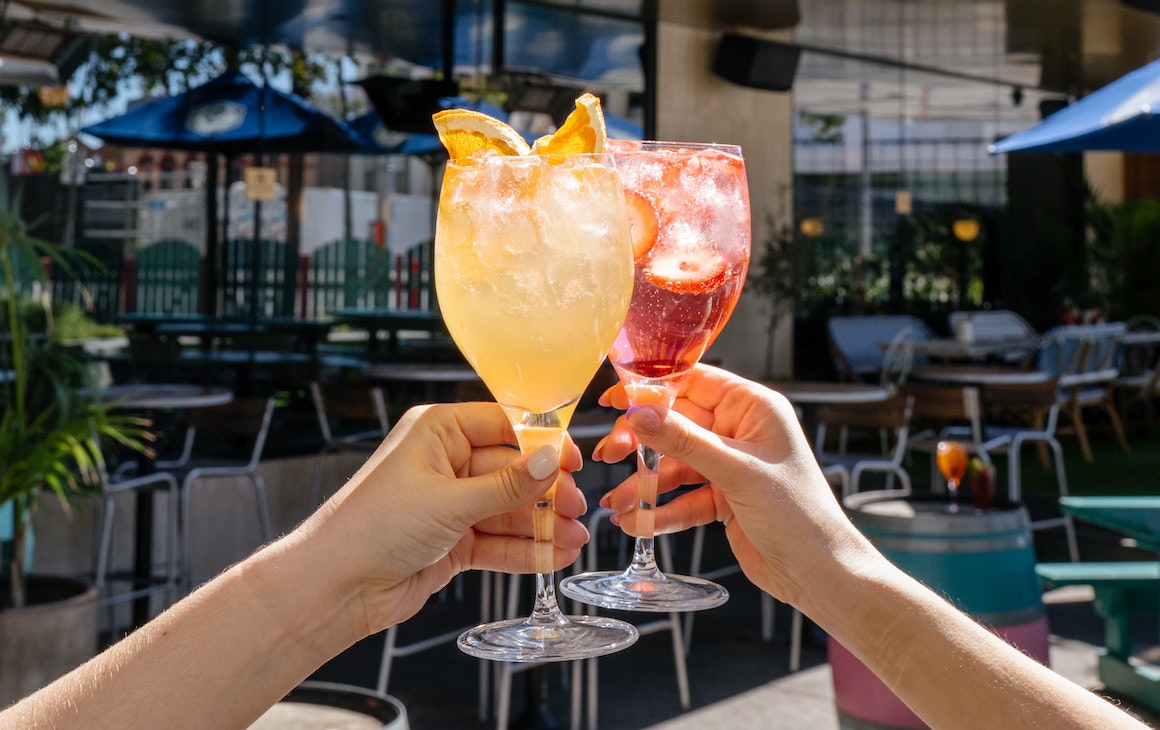 Two hands hold glasses of different coloured spritzes, one orange and one red.