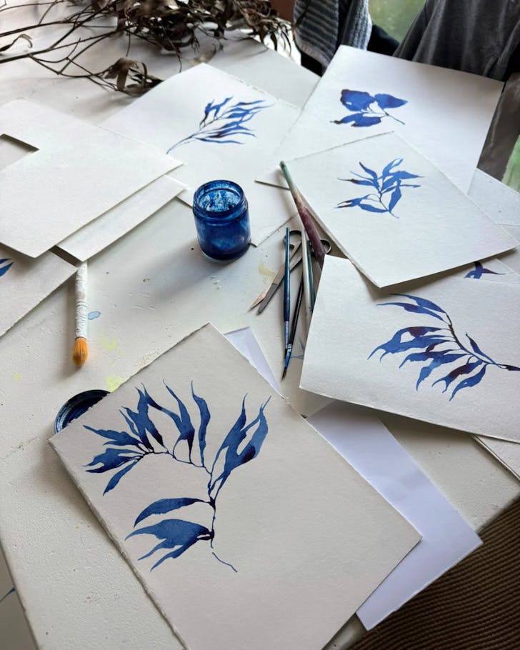 A series of blue watercolour flowers/plants painted on card, spread across a table with painting tools.