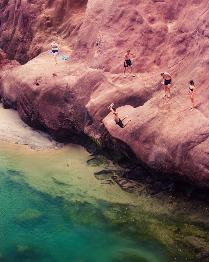 People jump from a striking red cliffside into green waters.