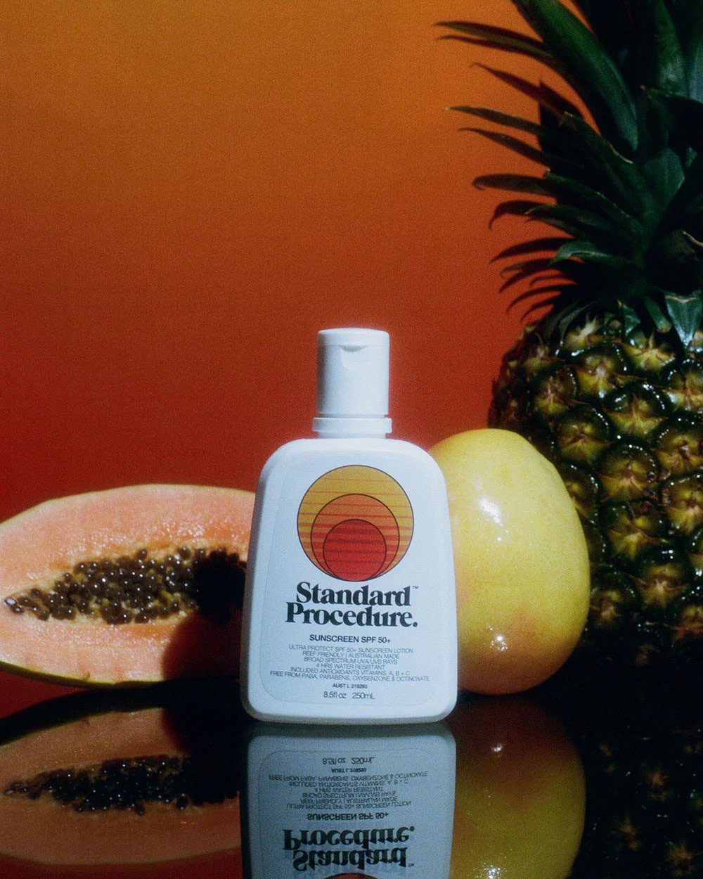 Une bouteille blanche de crème solaire Standard Procedure devant une papaye, une mangue et un ananas, sur fond orange. 