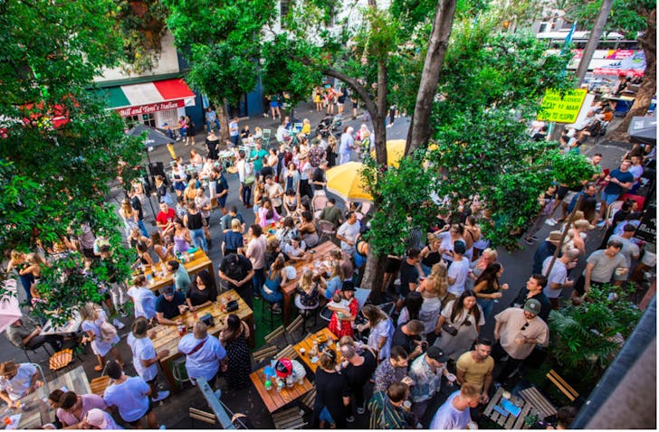 A wide shot from bird's eye view of a large crowd of people sitting outdoors at tables and walking around. People are chatting and socialising, the weather is nice and trees are around.