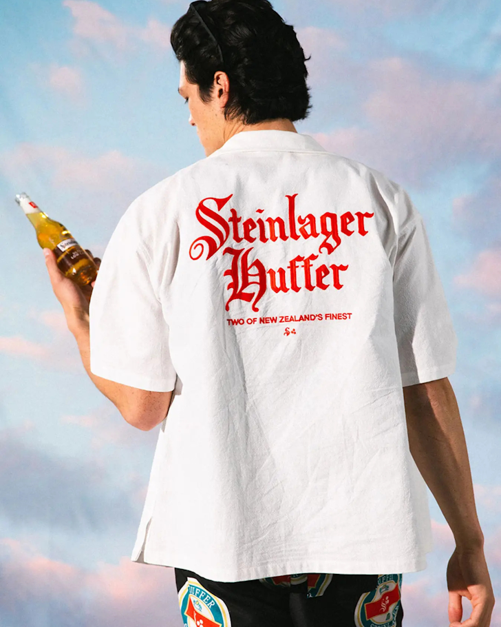 The back of someone drinking a Steinlager in front of a sunset with a white cubana shirt on with red Steinlager & Huffer calligraphy.