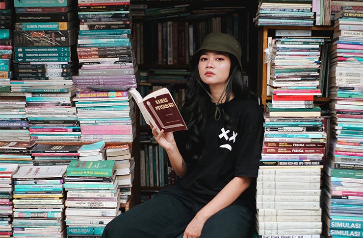 a woman wearing a hat holding a book and sitting between several large stacks of books