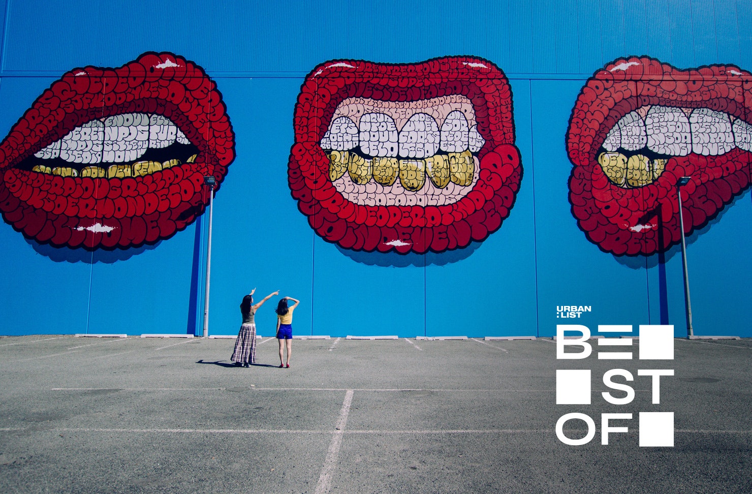 Two people gaze up at a huge street artwork showing three red mouths on a blue background.
