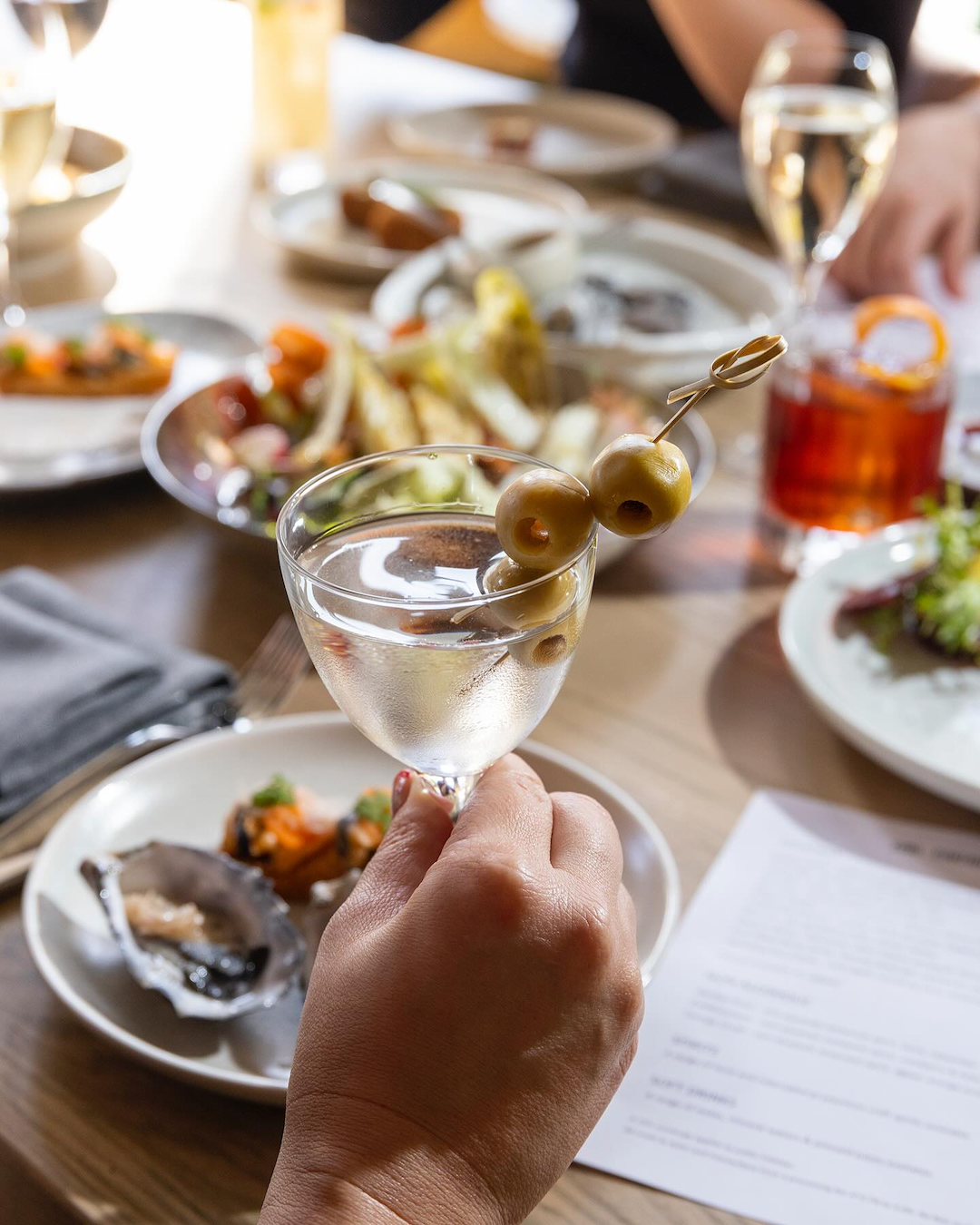 Someone holds a martini with olives over a table of bistro bites in Subiaco.