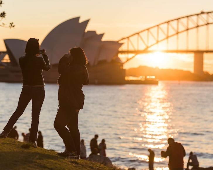 groups of people watch the sun set behind the sydney opera house and bridge from mrs macquarie's chair