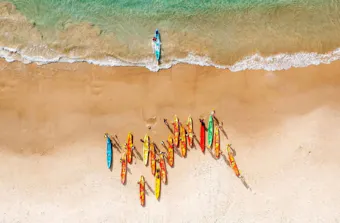 Aerial view of a sandy beach with a group of brightly coloured surf rescue boards lined up on the shore, while a single paddler approaches the beach from the clear turquoise water.