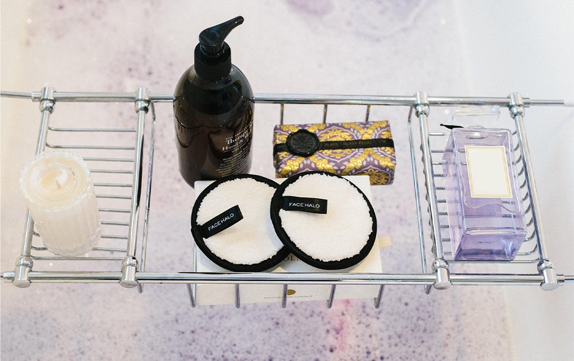 Several beauty products in a bath caddy over a bubbly bath.