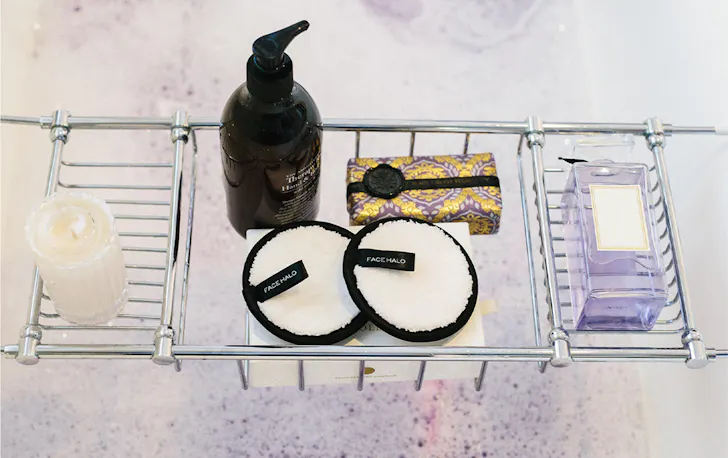 Several beauty products in a bath caddy over a bubbly bath.