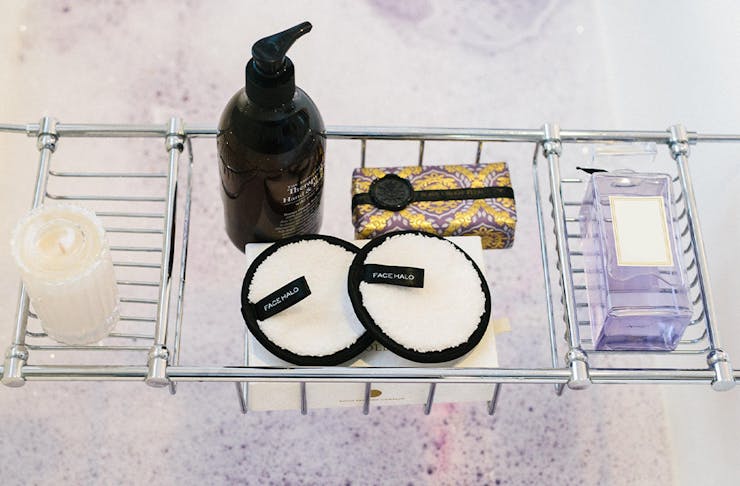 Several beauty products in a bath caddy over a bubbly bath.