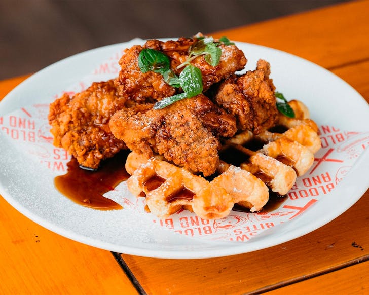 Waffles and crispy fried chicken with maple syrup on a white plate at Sweat Shop and Brew Kitchenâs bottomless fried chicken and waffles.