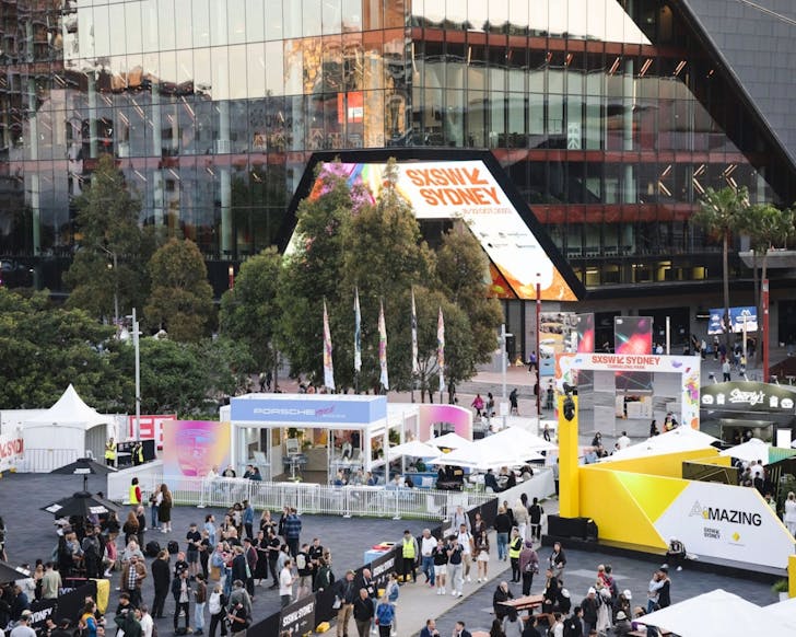 A birds-eye view wide shot of an outdoor SXSW event from 2024. In front of a large modern building, there is a large SXSW sign and people walking around the area enjoying the festival activities. 