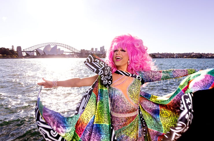 A person with bright pink hair and a beautiful rainbow coloured dress; the Sydney Harbour Bridge and Opera House are in the background. 