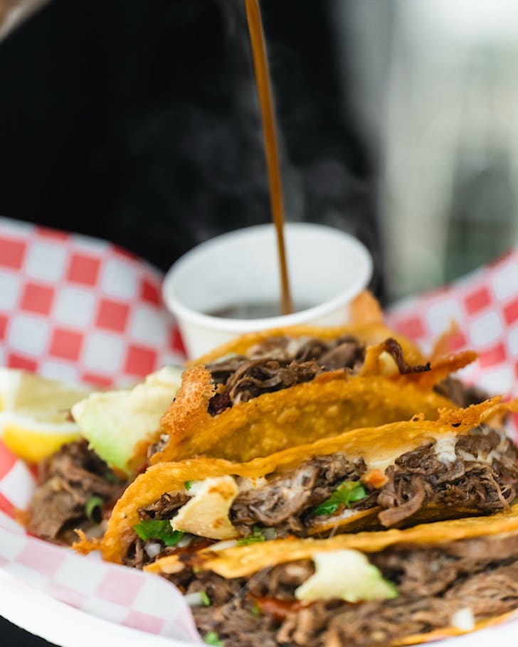Sauce is poured into a pottle beside a stack of birria tacos at Taco's Land.