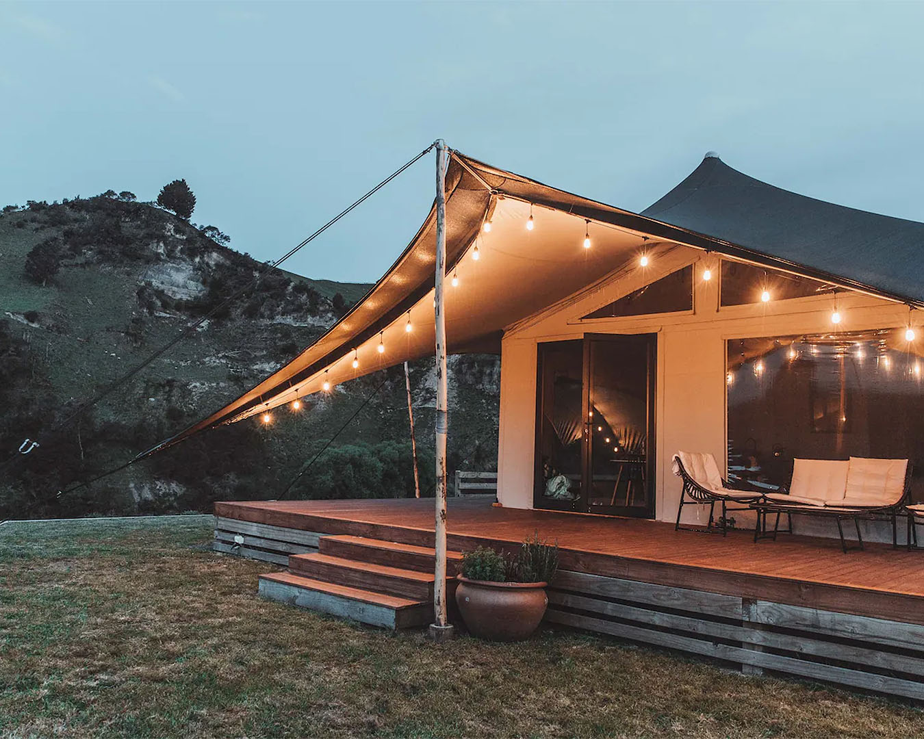 An a-frame tent is lit by fairy lights in the evening, and surrounded by a wooden deck. 
