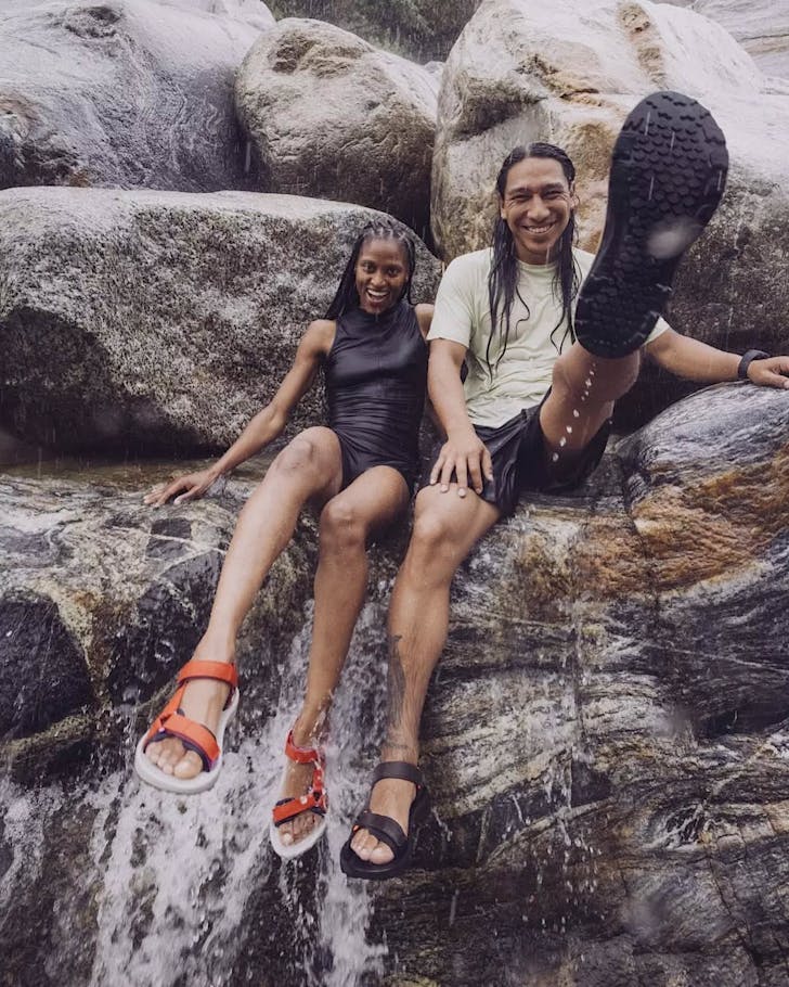 Two people sit in a waterfall while wearing Tevas. 