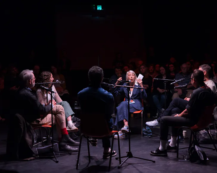 A panel of speakers sits in the round during an intimate Festival of Dangerous Ideas discussion, surrounded by audience members in a dark theatre setting.