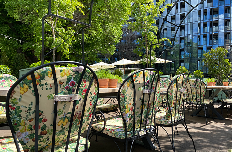 A long table at an outdoor bar underneath lush, green trees. A skyscraper towers in the background.