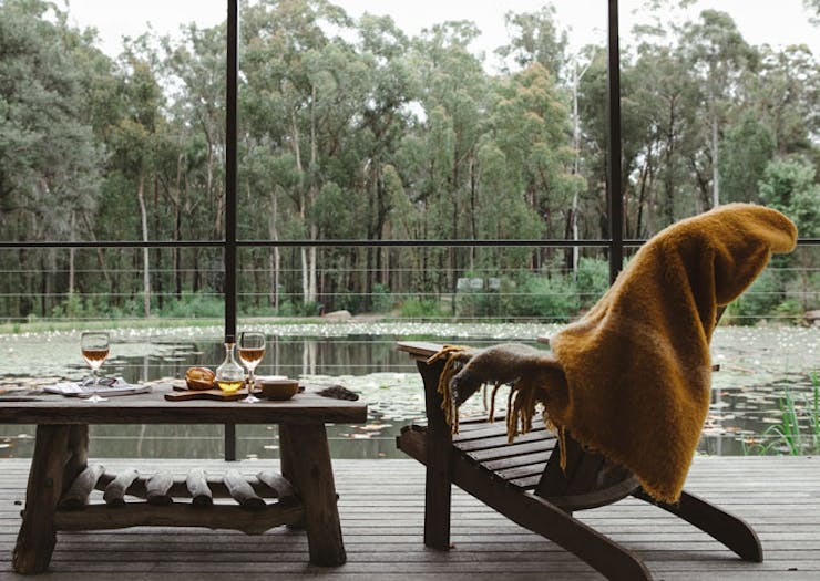 A yellow throw blanket over a deck chair, with wine and cheese laid out on a table, and a view over a lake. 