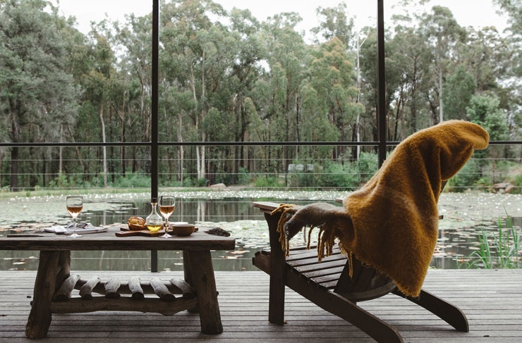 A yellow throw blanket over a deck chair, with wine and cheese laid out on a table, and a view over a lake. 