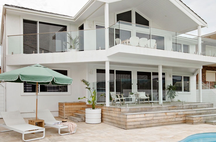 A large white beach house, with a pool in the foreground, deck chairs and a green umbrella. 