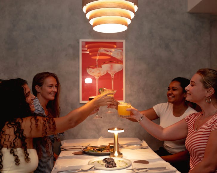 A group of four women sit in a dimly lit restaurant booth toasting their glasses while smiling. Dishes of food sit on the table between them and the space is artfully decorated.
