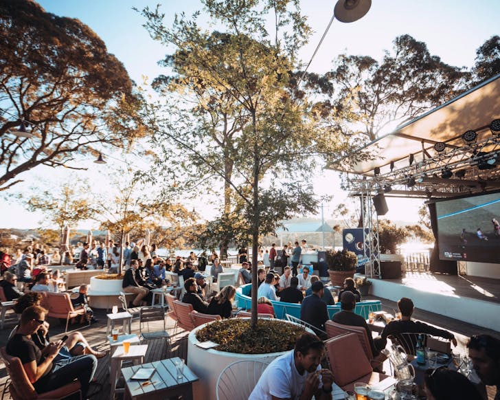 A far wide shot of The Newport exterior seating, with a stage and large screen just visible to the left and the ocean in the background. The setting is daytime and their are many people sitting on the outdoor seating. 