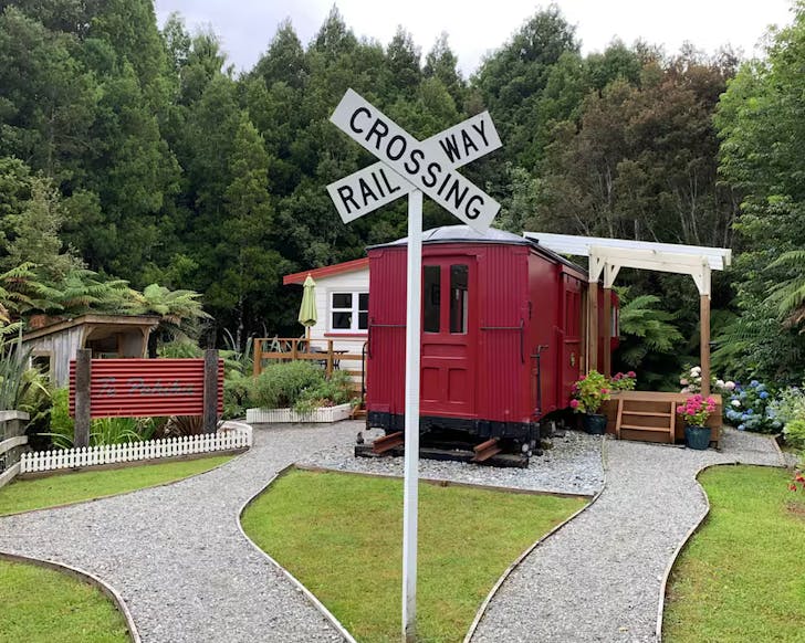 A red train carriage converted to accommodation sits before native bush, with neatly manicured lawns out front and a traditional railway crossing X sign.