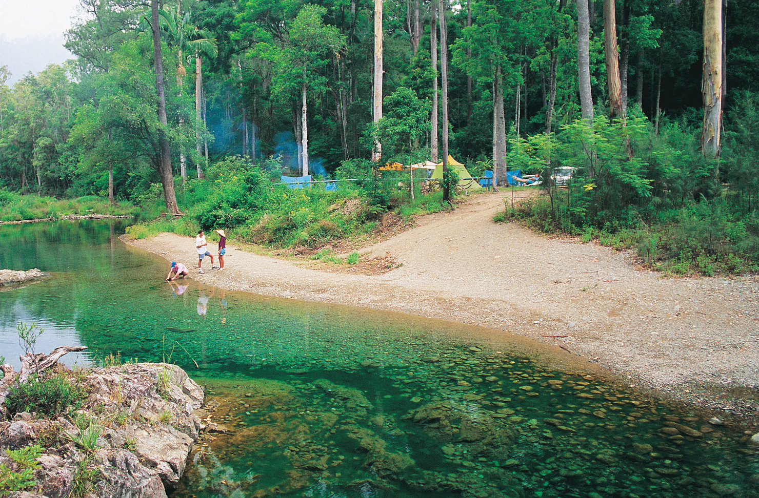 Three campers gather by a creek between their campsites and the water.