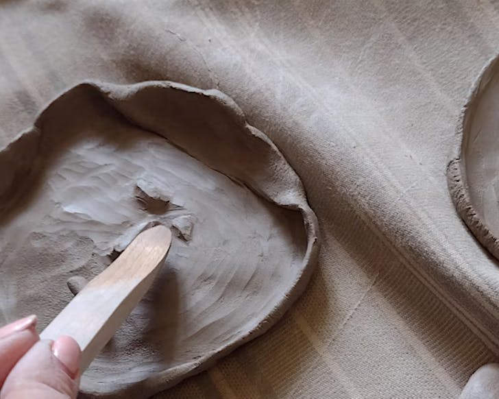 Someone puts the finishing touches on a drying clay trinket dish with a wooden tool.