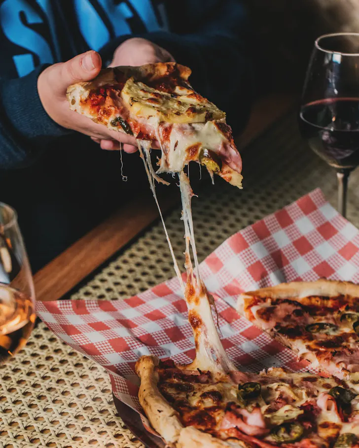 Hand lifting a slice of woodfired pizza with stretchy melted cheese over a whole pizza on a red gingham-lined tray, served alongside glasses of wine.