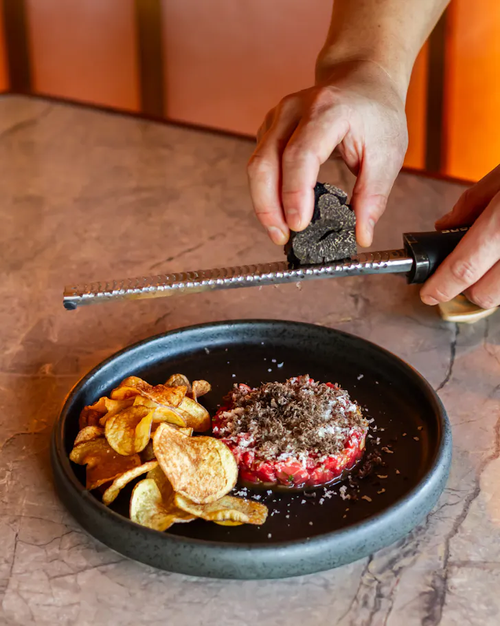 A chef shaves fresh black truffle over a neatly plated wagyu beef tartare topped with grated cheese, served alongside crisp house-made potato chips on a dark ceramic plate.