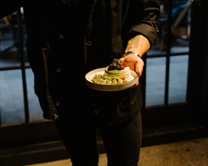 Yovin is dressed in black and holds out a plate of perfect matcha mousse pavlova